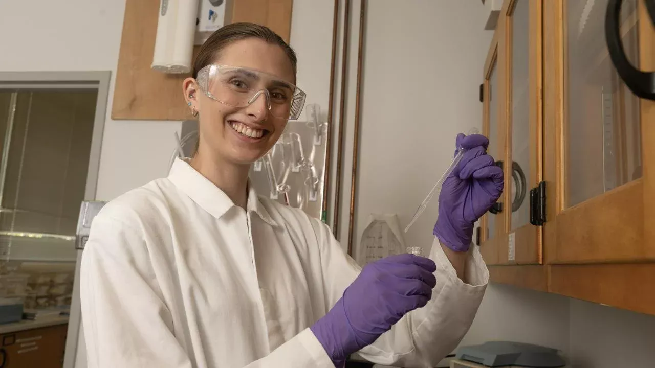 Brook wears a lab coat, gloves and safety glasses while holding a test tube